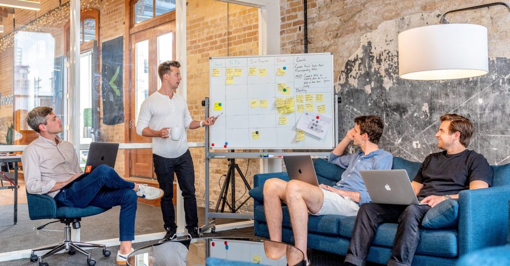 three men sitting while using laptops and watching man beside whiteboard