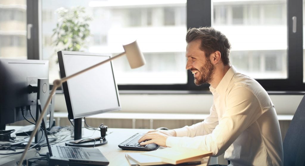 Man in white dress shirt sitting on black rolling chair while facing black computer set and smiling